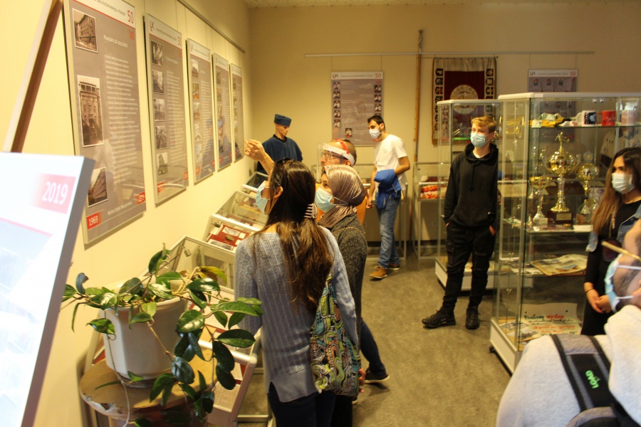 Zagraniczni studenci medycyny w pomieszczeniu w którym znajdują się pamiątki dokumentujące 50 lat historii Uniwersytetu Jana Kochanowskiego. Na ścianie tablice pamiątkowe, w środku gabloty z pucharami.