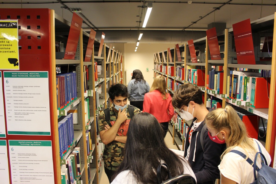 Studenci sekcji English Division kierunku lekarskiego UJK przeglądają książki pomiędzy regałami biblioteki. Mają założone maseczki.