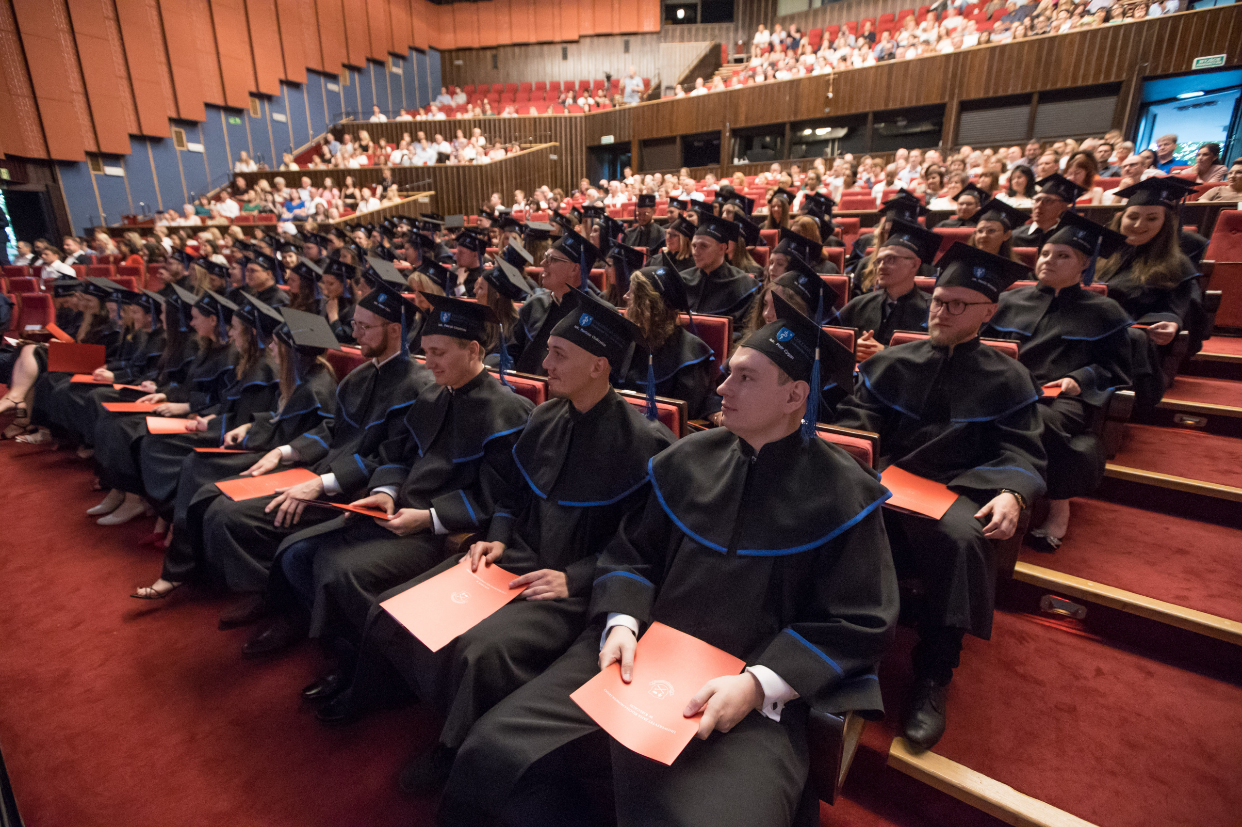 Zbiorowe zdjęcie absolwentów kierunku lekarskiego. Wszyscy siedzą w togach na sali KCK-u, trzymają dyplomy.