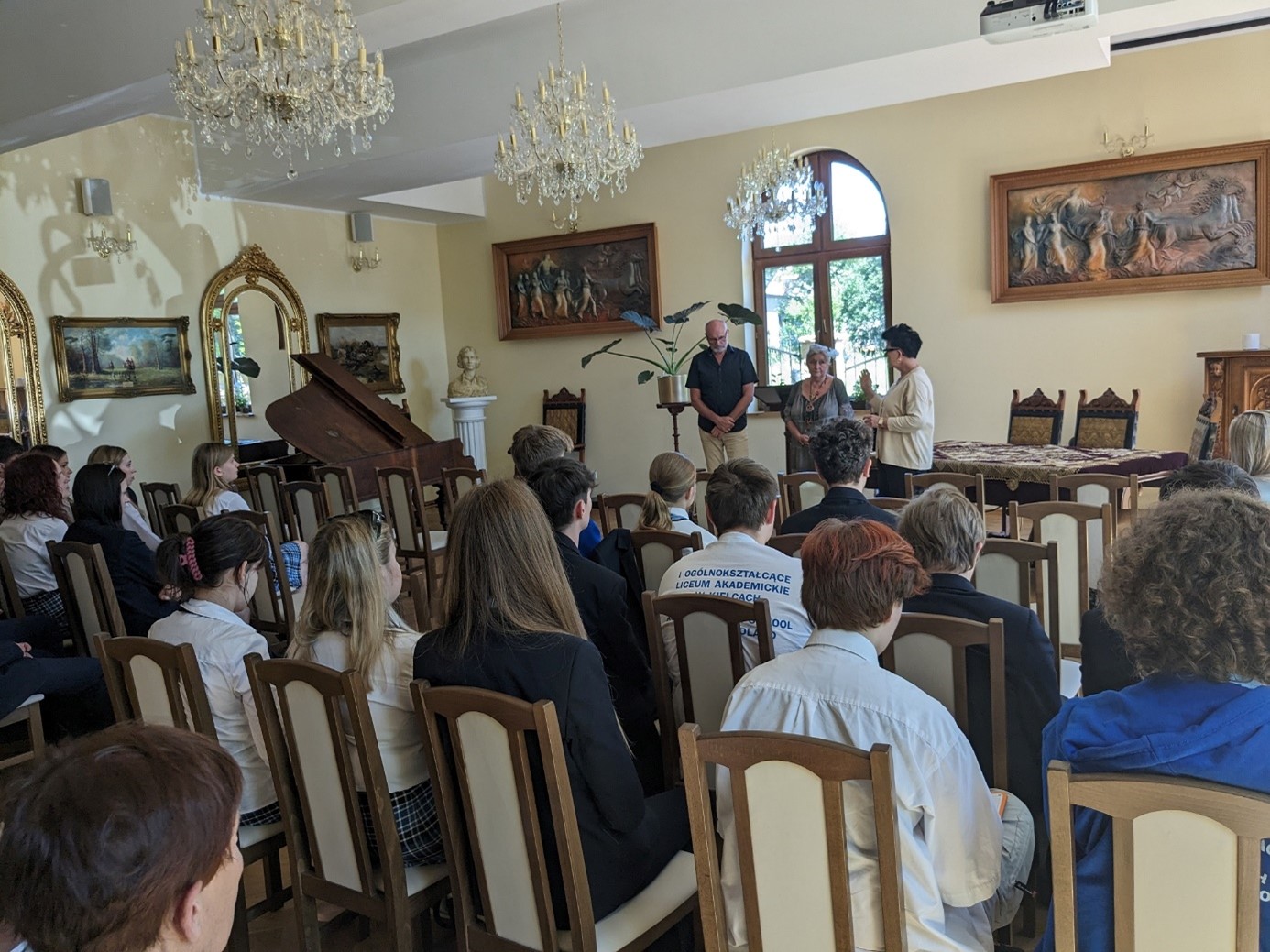 Uczniowie I Ogólnokształcącego Liceum Akademickiego im. Janiny Kossakowskiej-Dębickiej w Kielcach siedzą na krzesełkach w sali szkoły. Prof. UJK dr hab. Mariola Wojciechowska rozmawia z dwojgiem przedstawicieli kadry szkoły.