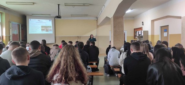 przedstawicielka kierunku Turystyka prozdrowotna wyświetla prezentację i opowiada o kierunku. Na dużej sali uczniowie siedzą na ławkach i słuchają.