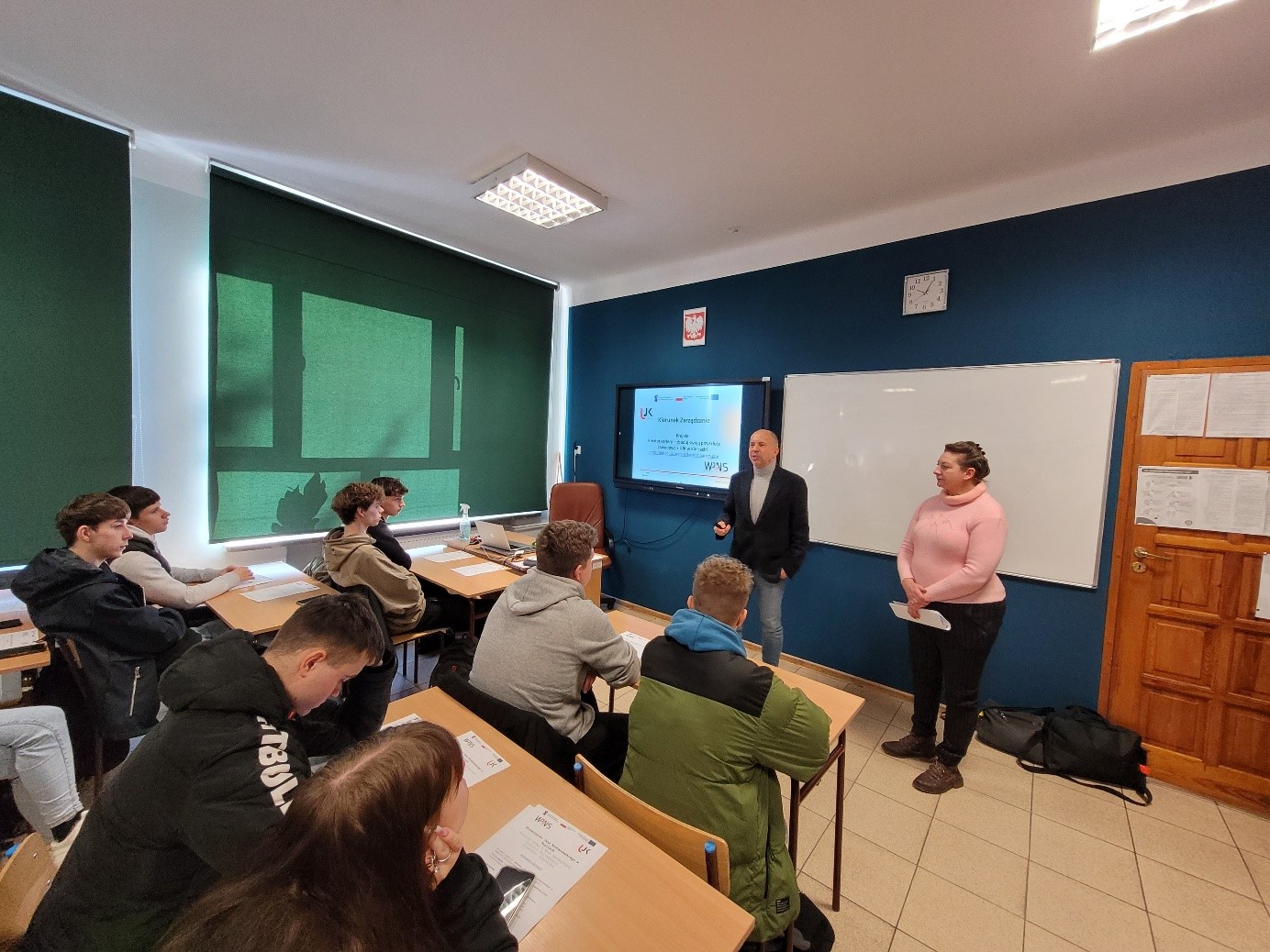 Przedstawiciele kadry UJK prezentują w jednej ze szkół zakres wsparcia w ramach projektu na kierunku Zarządzanie. W klasie siedzą uczniowie w ławkach.