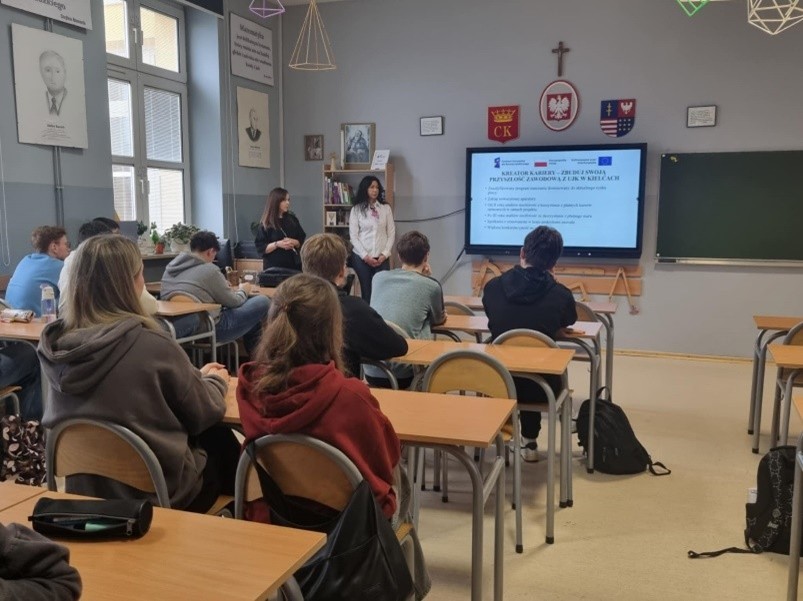 Przedstawicielki kadry UJK prezentują w jednej ze szkół zakres wsparcia w ramach projektu. W klasie siedzą uczniowie w ławkach.