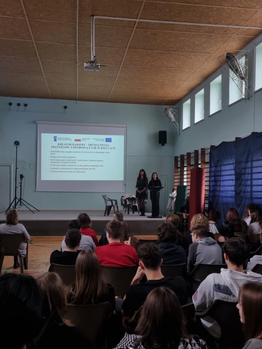 Przedstawicielki kadry UJK w Kielcach przedstawiają na prezentacji wyświetlanej na sali gimnastycznej ofertę kierunku Kosmetologia. Uczniowie siedzą na krzesełkach w rzędach.