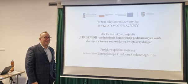 Na zdjęciu widać mężczyznę - prowadzącego wykład motywacyjny, który stoi obok ekranu projekcyjnego, na którym wyświetlana jest prezentacja.