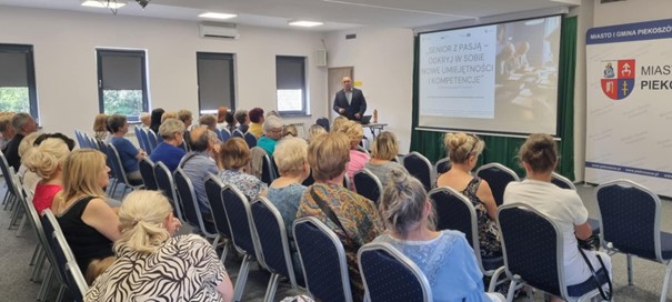 Na zdjęciu widać salę wykładową, prowadzącego wykład mężczyznę. Na sali widoczni są Kobiety i Mężczyźni, którzy słuchają wykładu i patrzą na wyświetlaną prezentację.