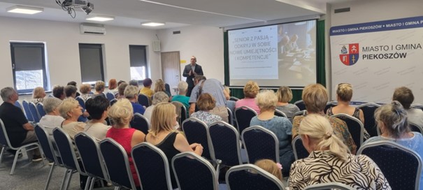 Na zdjęciu widać salę wykładową, prowadzącego wykład mężczyznę. Na sali widoczni są Kobiety i Mężczyźni, którzy słuchają wykładu i patrzą na wyświetlaną prezentację.