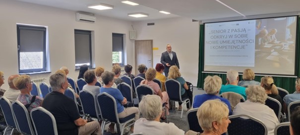 Na zdjęciu widać salę wykładową, prowadzącego wykład mężczyznę. Na sali widoczni są Kobiety i Mężczyźni, którzy słuchają wykładu i patrzą na wyświetlaną prezentację.