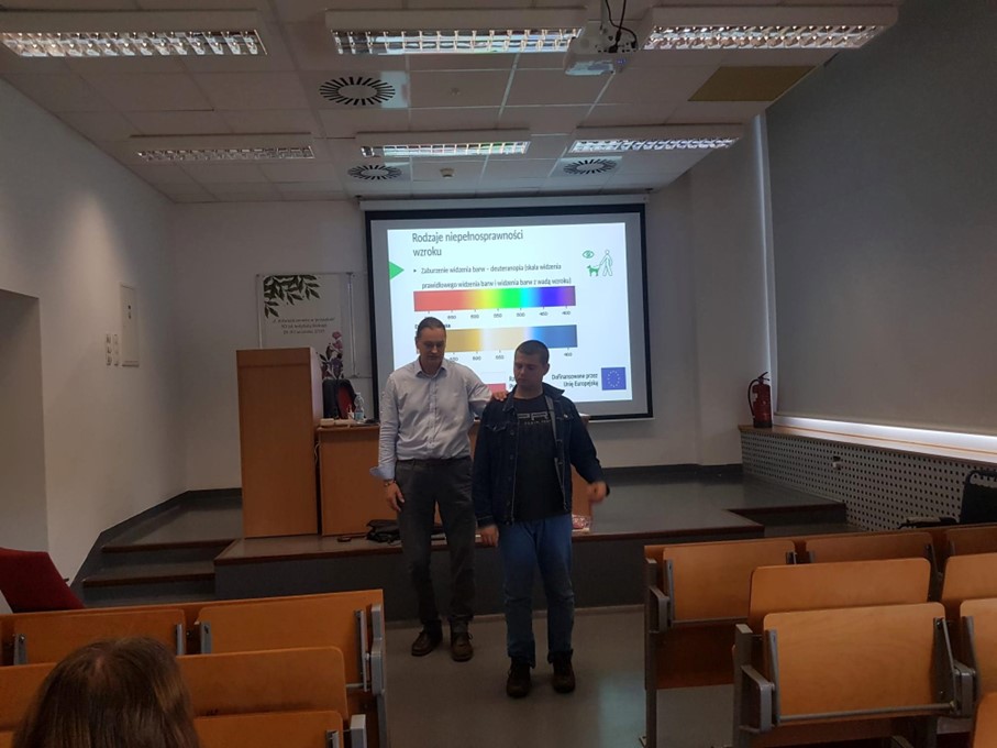 na zdjęciu widać salę wykładową, w tle ekran projekcyjny z wyświetloną prezentacją szkoleniowa. Na środku sali widać trenera i jednego z Uczestników, którzy wykonują ćwiczenie.