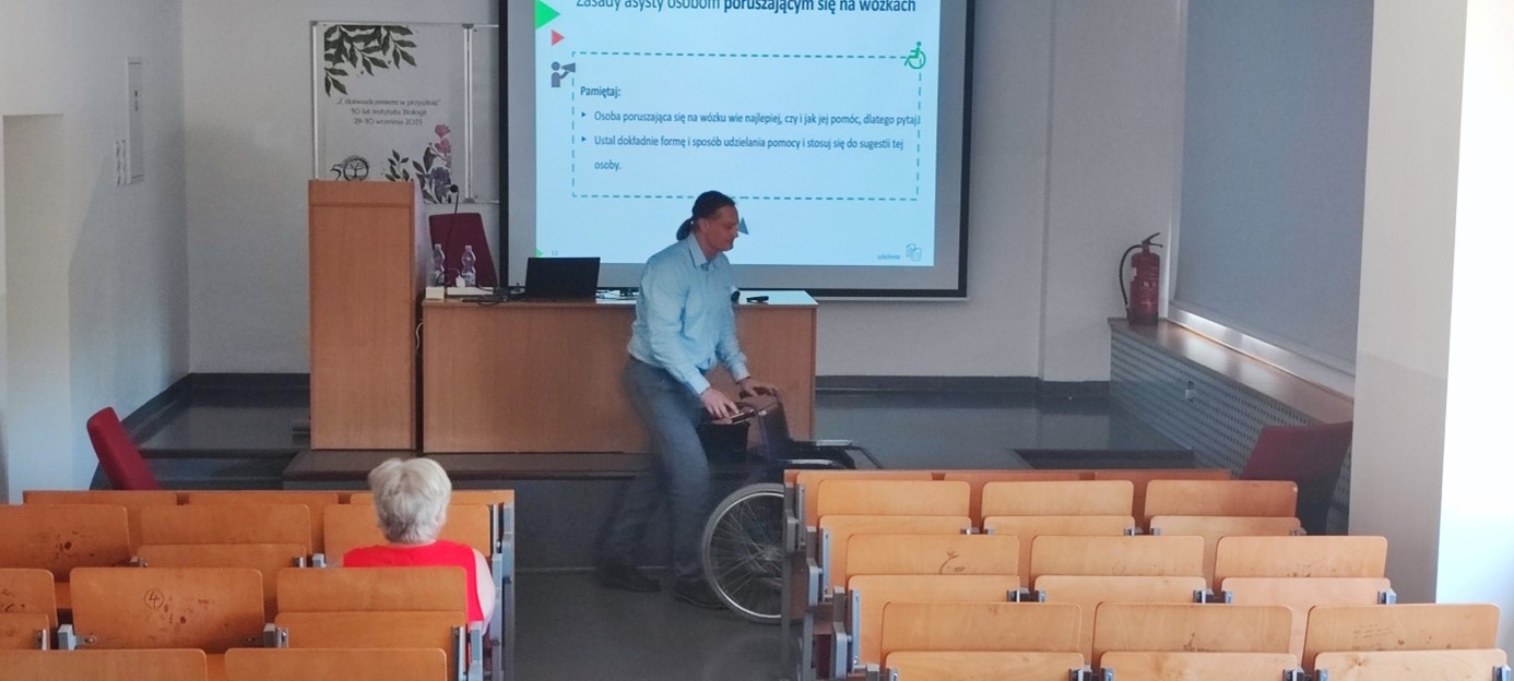 Na zdjęciu widoczna jest sala dydaktyczna. Trener trzyma wózek inwalidzki.